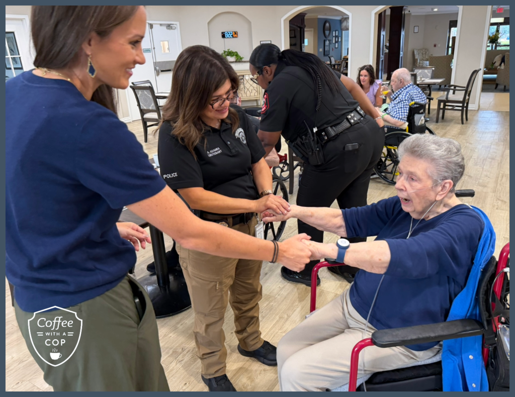 A Meadows Place police lieutenant and another attendee hold hands with a smiling resident in a wheelchair during Coffee with a Cop at The Jasper of Meadows Place.