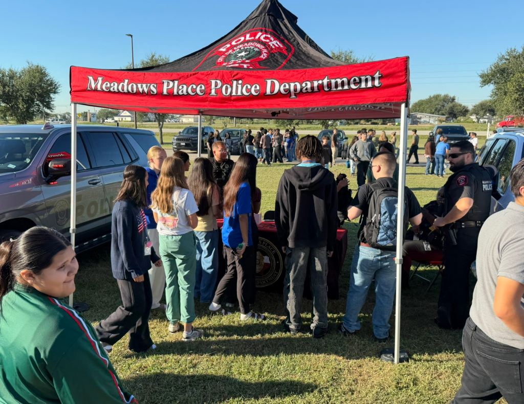 Students gather around the Meadows Place Police Department tent at Needville High School's Law Enforcement & Public Service Showcase. Officers speak with students beside police vehicles while other groups visit displays in the background.