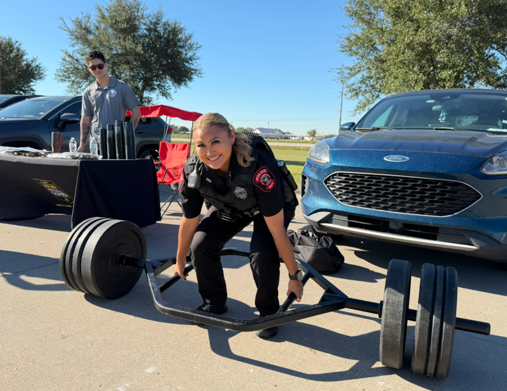 A smiling Meadows Place police officer poses in a lifting position with a heavily weighted trap bar during an outdoor law enforcement event. A man stands near a display table behind her, with parked vehicles in the background.