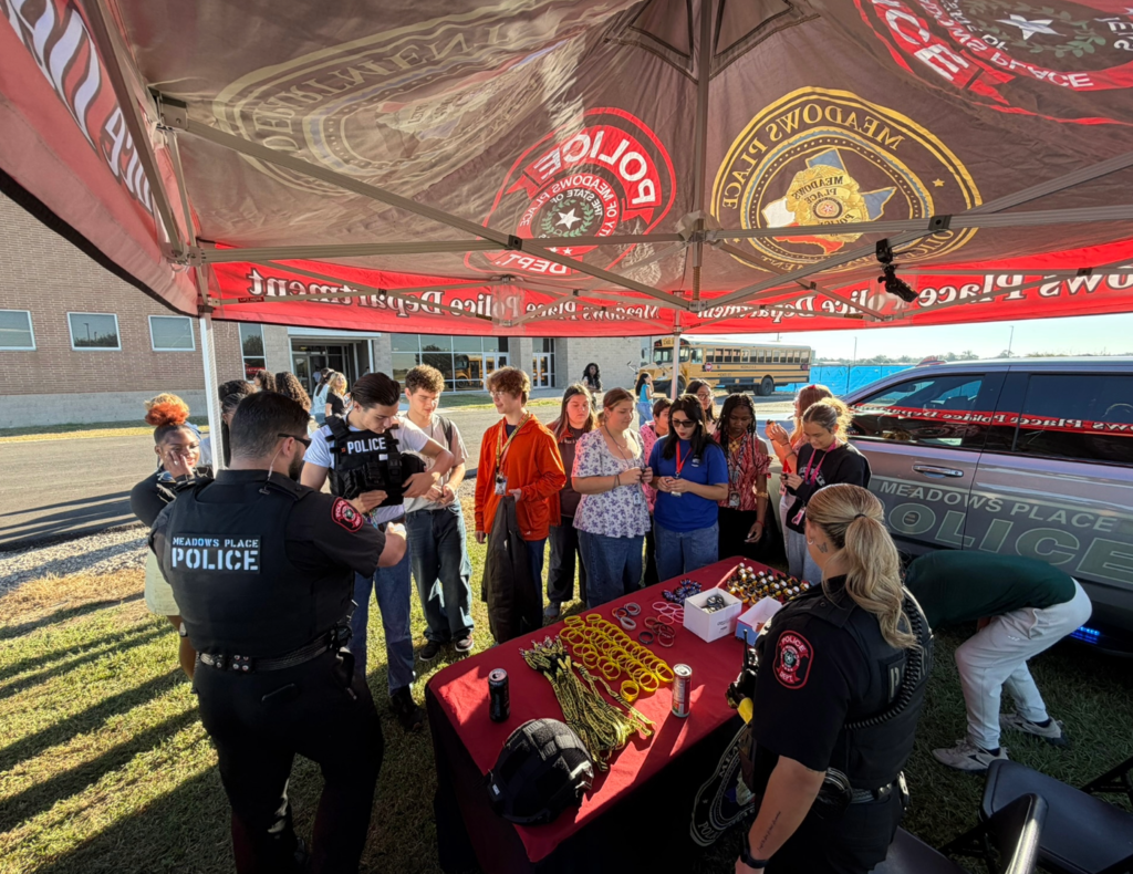 Meadows Place police officers interact with students at Needville High School's Law Enforcement & Public Service Showcase. One student tries on a police tactical vest while others gather around a table with giveaways and equipment under a Meadows Place Police Department tent.