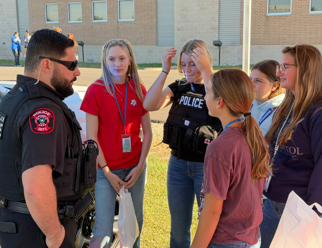 A Meadows Place police officer speaks with a group of five students during an outdoor law enforcement event. One student wears a police tactical vest while the others gather around and listen.