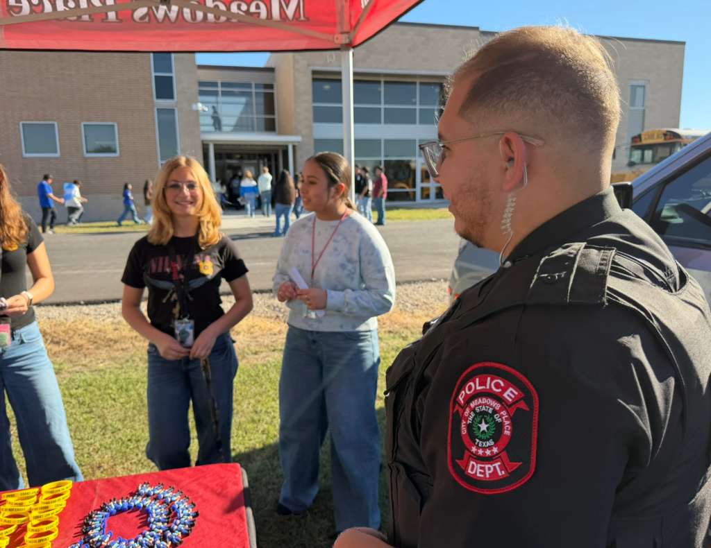 A Meadows Place police officer speaks with students at an outdoor law enforcement event. A table displaying colorful giveaways and yellow wristbands sits beneath the department's red canopy, with a school building in the background.