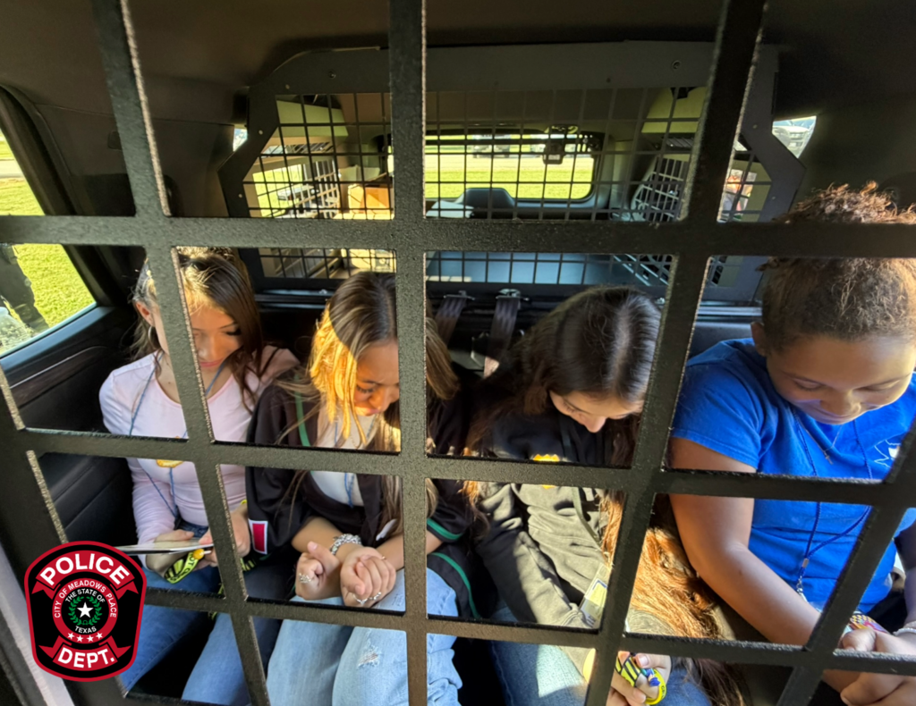Four students sit together in the back seat of a Meadows Place police vehicle, viewed through the metal security partition separating the front and rear seats.