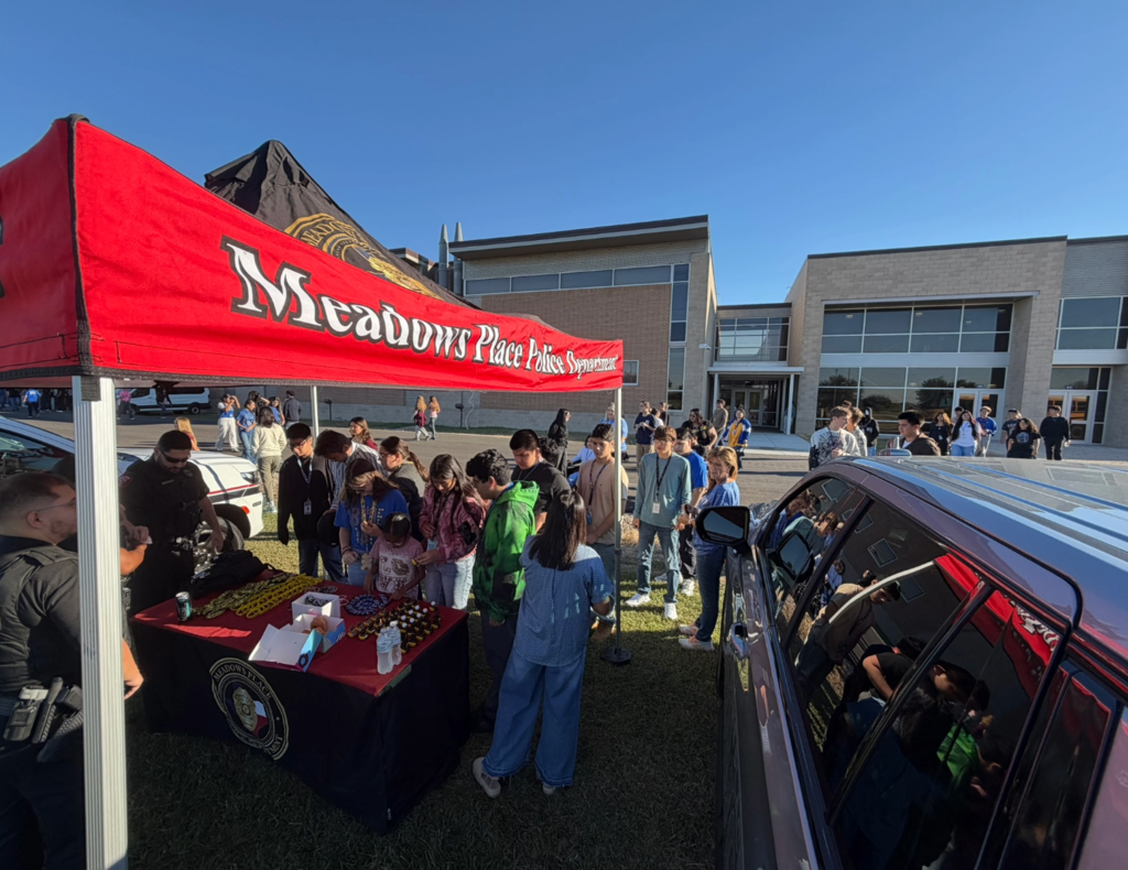 Students gather around the Meadows Place Police Department's booth at a high school law enforcement event. Officers interact with students beneath the department's red canopy, with a police SUV and school building in the background.