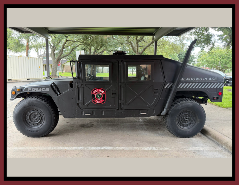 **Alt text:** Side view of a black Meadows Place Police Department rescue Hummer parked under a covered structure. The vehicle displays Meadows Place Police markings on the doors and sides and appears freshly restored with a matte black finish.