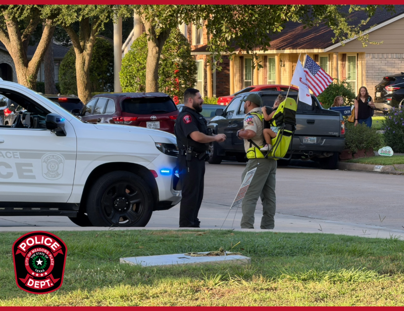 Meadows Place police officer talks with an adult carrying a child near a police vehicle in a residential neighborhood.