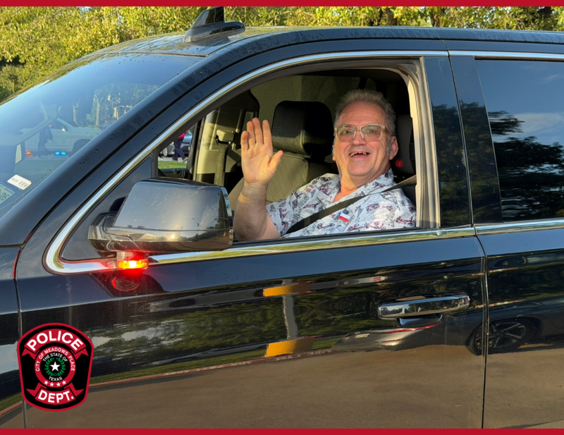 Smiling man waves from the driver’s seat of a vehicle during first-day-of-school activities.