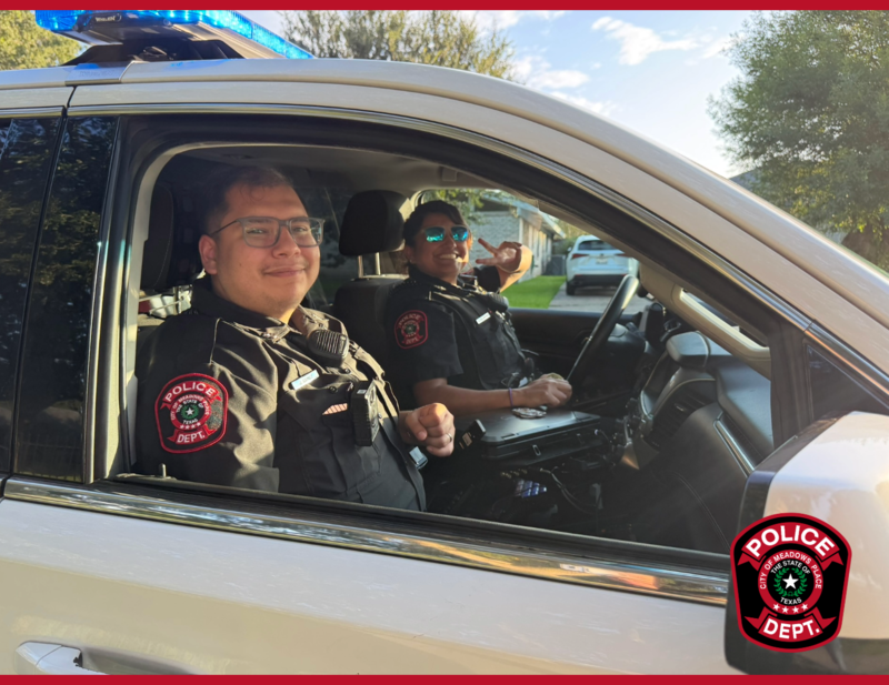 Two Meadows Place police officers smile from inside a police vehicle, with one officer giving a peace sign.