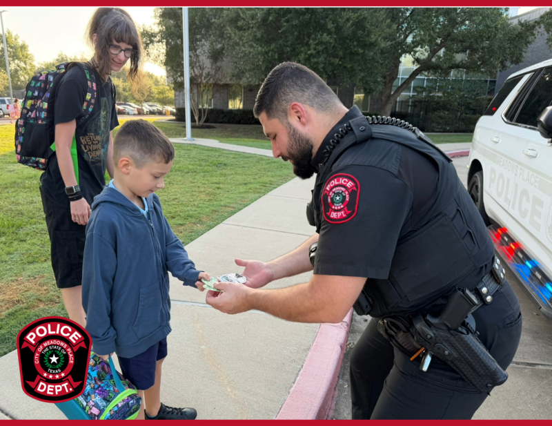 Meadows Place police officer handing items to a young student outside a school as an adult stands nearby.