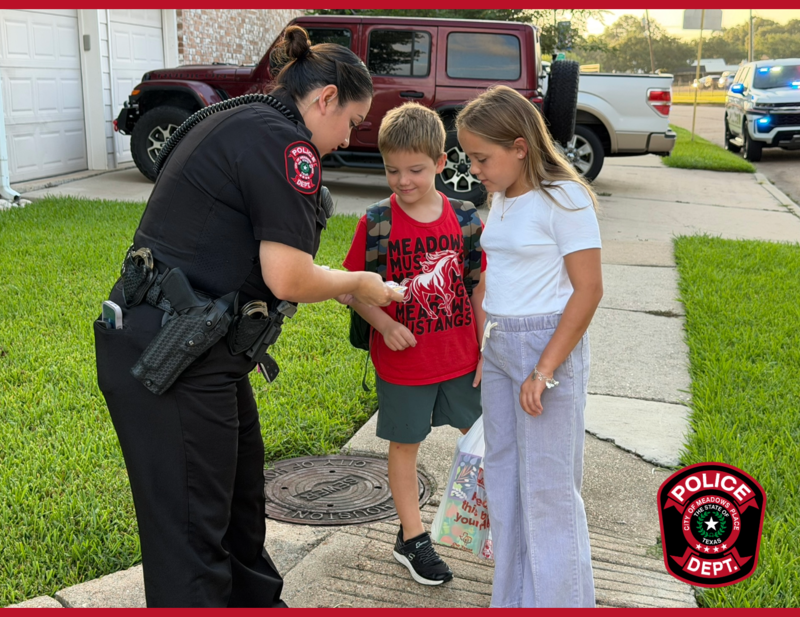 Meadows Place police officer hands items to two students with backpacks on a neighborhood sidewalk before school.