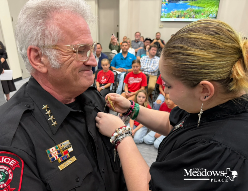 Meadows Place Police Chief Jack Ashton’s daughter pins his badge during his swearing in ceremony as family, friends, officers, and community members look on. The City of Meadows Place logo appears in the lower right corner.