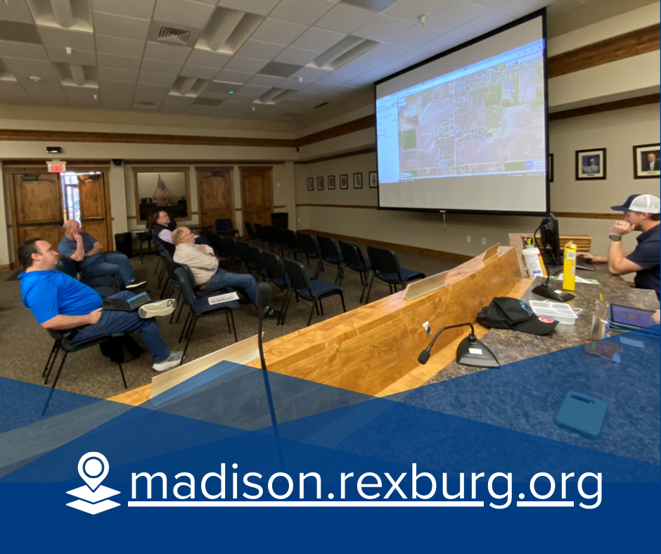 people sitting in council chambers facing a projector screen showing an image of mapping software. one more person is seen behind a counter running the computer