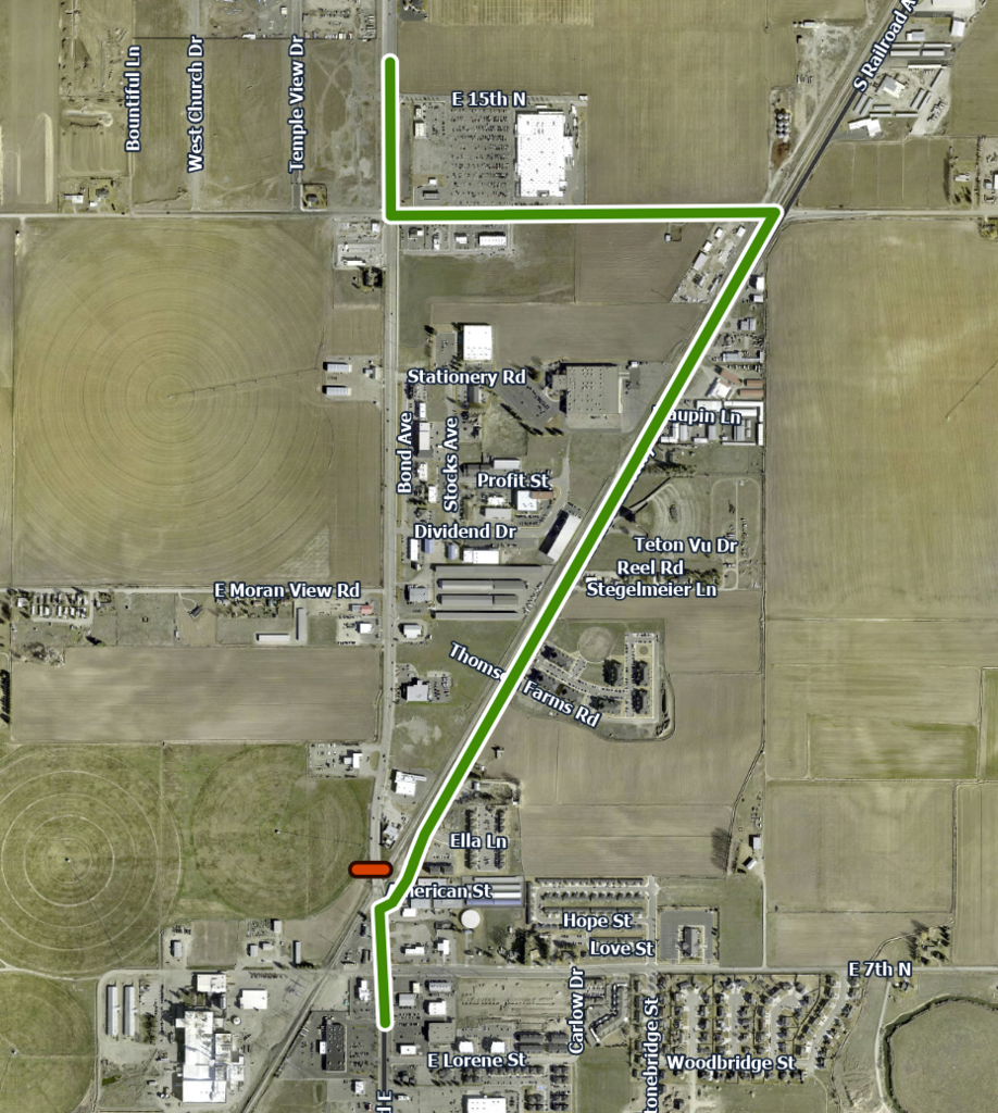 Aerial view with a green line indicating a route, intersecting a road with a red dot. Industrial buildings and open fields surround the area.
