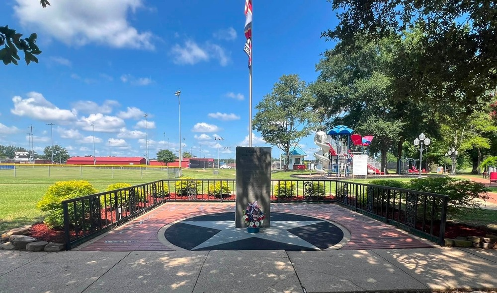 A veterans memorial stands beneath a U.S. flag in a landscaped park, featuring a black granite monument, a patriotic wreath, and a star-shaped brick plaza surrounded by decorative fencing. In the background are Richland's baseball fields, a colorful playground, walking paths, and large shade trees under a bright blue sky.