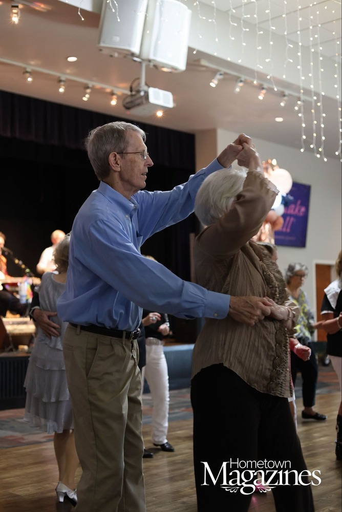 An older couple dances together on the dance floor during a community event at the Richland Senior Center. The man in a blue button-down shirt twirls his partner beneath string lights while other couples dance in the background. A live band performs on stage, creating a festive atmosphere during the Snowflake Ball