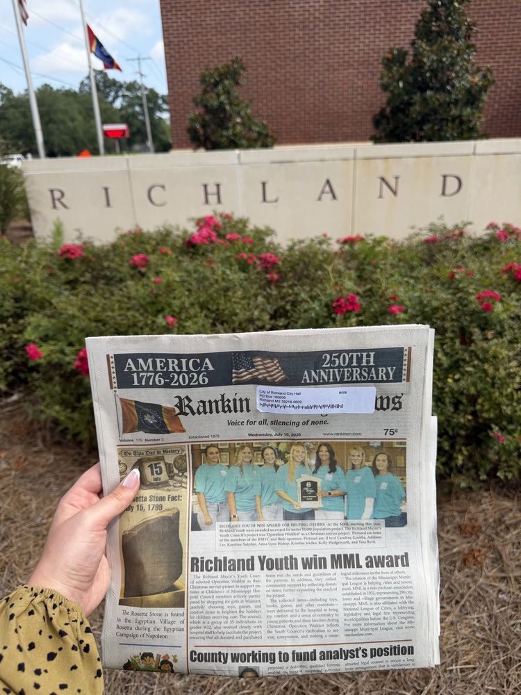 A person holds a copy of the Rankin County News in front of the City of Richland entrance sign and landscaped flower beds. The newspaper's front-page headline reads, "Richland Youth win MML award," highlighting the Richland Mayor's Youth Council's recognition for its community service project. City Hall and flags are visible in the background