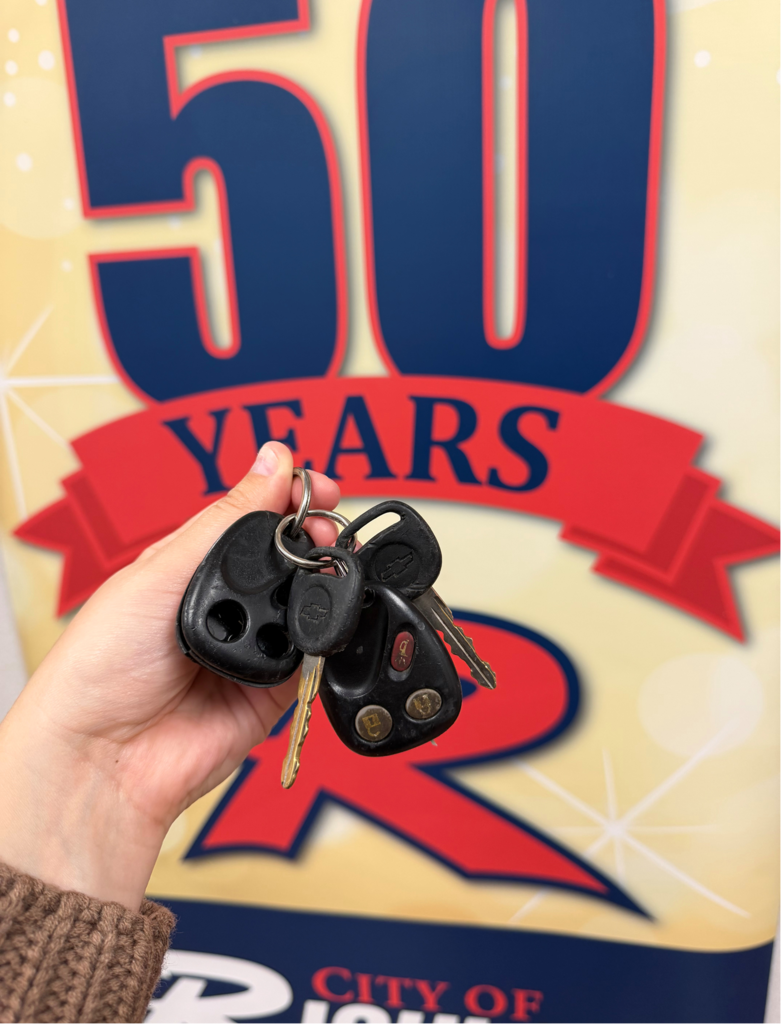 A hand holds several car keys in front of a "50 Years" anniversary sign with the letter "R" in the background.