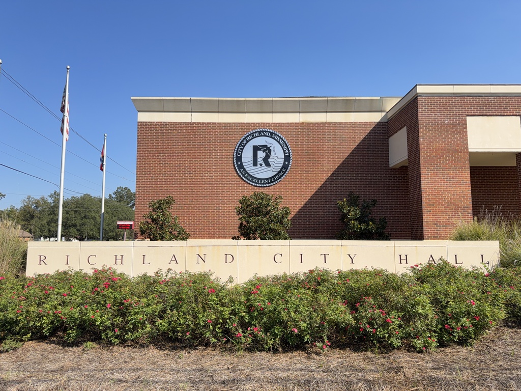 Exterior view of Richland City Hall featuring the newly installed City of Richland emblem on the brick building, with the βRichland City Hallβ sign, landscaped flower beds, and American and Mississippi flags visible under a clear blue sky.