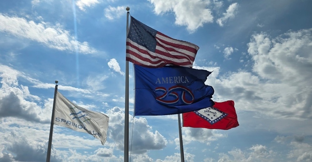 A wide-angle outdoor photograph showing four flags flying on tall flagpoles against a dramatic summer sky filled with large white clouds and bright sunlight.  Centered in the image is a tall flagpole displaying the United States flag at the highest position. The American flag is fully extended by the wind, with its red and white stripes rippling across the sky and its blue field of stars visible near the flagpole. Sunlight filters through the clouds behind it, creating bright beams of light and emphasizing the flag as the focal point of the photograph.  Directly beneath the American flag on the same pole is a blue America 250 commemorative flag. The flag displays the text “America 250” in white lettering with a red, white, and blue stylized “250” logo. The flag is also billowing strongly in the wind, symbolizing the nationwide celebration of the 250th anniversary of the United States.  To the left, a separate flagpole flies the white City of Springdale flag. The flag features the city’s logo and the word “Springdale” in blue lettering. The wind causes the fabric to stretch outward, revealing portions of the logo and text.  To the right, another flagpole displays the Arkansas state flag. The bright red flag features a large white diamond outlined in blue with stars and the word “Arkansas” in the center. Like the other flags, it is extended by the breeze and stands out against the sky.  The lower portion of the image shows a tree-lined landscape with dense green foliage stretching across the horizon. No people are visible. The photograph conveys a sense of civic pride, patriotism, and community identity, with the American, America 250, Arkansas, and Springdale flags flying together beneath a bright blue sky and towering clouds.