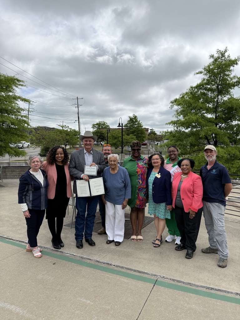 A group of ten people stand together in a loose line on a wide concrete walkway outdoors, smiling and facing the camera as if posing for a commemorative photo. The sky above them is filled with thick gray clouds, creating a soft, diffused light. In the background are leafy green trees, a black metal railing, decorative streetlamps, and utility poles with visible power lines, suggesting a landscaped public space such as a park, trail, or riverside overlook.  At the center of the group, a person wearing a light-colored cowboy hat, gray blazer, button-down shirt, and jeans holds open a framed document or proclamation, angling it toward the camera so the pages are visible. The document appears formal, with printed text and signatures. Standing closely beside them is another person wearing a blue long-sleeve top and white pants, smiling and leaning slightly toward the center.  To the left side of the group, one person wears glasses and a navy jacket layered over a striped top, standing with hands relaxed at their sides. Next to them, another person wears a black dress paired with a pink blazer, standing confidently with a slight forward lean. Slightly behind the central figures, a person with facial hair and sunglasses resting on their head peeks between shoulders, partially visible but smiling.  To the right of the central pair, several individuals stand closely together in a cohesive cluster. One wears a bright green outfit with a bold, multicolored patterned top, standing with feet slightly apart. Another wears a navy blazer with a name badge pinned to it over a patterned dress, hands clasped in front. Nearby, a person in a coral blazer over a dark outfit stands with a relaxed, upright posture. Another person in a green top and light-colored sneakers stands slightly behind the front line, smiling.  At the far right edge of the group, a person in a baseball cap, navy polo shirt, and light gray pants stands slightly apart with a casual stance, hands resting near their sides.  Overall, the group’s body language and the prominently displayed document suggest a formal recognition, announcement, or community celebration taking place in a public outdoor setting.