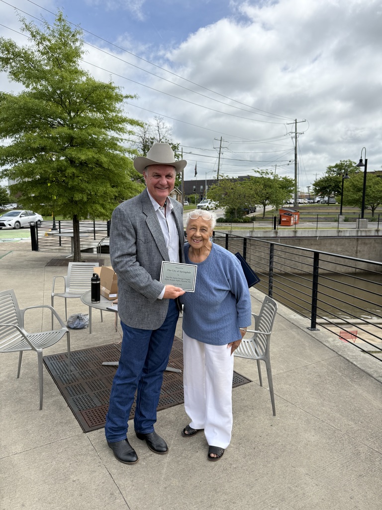 Two people stand close together on a paved outdoor walkway, smiling warmly at the camera while holding a small plaque between them. The setting appears to be a landscaped public space, possibly a park or riverside area, with a black metal railing running along the right side and a shallow waterway or channel below.  The person on the left wears a light-colored cowboy hat, a gray textured blazer over a button-down shirt, blue jeans, and dark boots. They stand angled slightly toward the other person, holding the plaque with both hands at chest height. The plaque is rectangular with a light background and dark border, featuring printed text that suggests a formal recognition or presentation.  The person on the right stands close at their side, leaning in slightly with a relaxed and cheerful posture. They wear a blue long-sleeve top with a subtle textured pattern, white pants, and dark sandals. A pair of glasses rests on top of their head. One arm is positioned near the plaque while the other rests naturally by their side, and they smile broadly.  Behind them, several lightweight metal chairs and a small round table are arranged on the concrete surface, suggesting a casual gathering or small event setup. On the table are a water bottle and a cardboard box. To the left, a leafy green tree provides a soft natural backdrop, while further back are additional trees, a street with parked cars, and utility poles with visible power lines stretching across the sky.  The sky is partly cloudy with patches of blue visible through large, soft clouds, casting even, natural light across the scene. The overall composition and the shared plaque suggest a moment of recognition, presentation, or celebration captured in a relaxed outdoor environment.