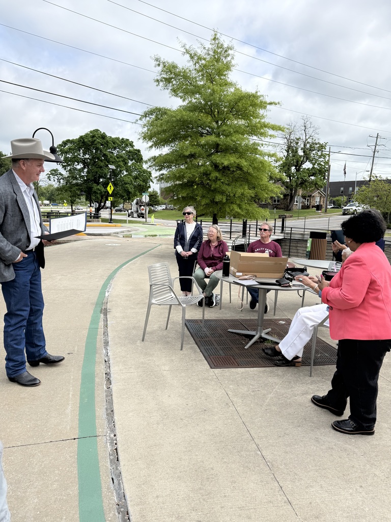 A small group gathers on a curved concrete walkway in an outdoor public space, appearing to take part in a casual ceremony or presentation. The sky is overcast with patches of lighter clouds, and the area is surrounded by green trees, a black metal railing, and nearby streets with parked cars and utility poles visible in the background.  On the left, a man wearing a light-colored cowboy hat, gray blazer, button-down shirt, jeans, and dark boots stands facing the group. He holds an open folder or binder at chest height and appears to be reading or speaking from it, his body angled slightly toward the seated audience.  To the right, several people are arranged around a small round metal table and lightweight metal chairs. Two women and one man sit at the table, while another woman stands just behind them. The seated individuals face the speaker, listening attentively. One woman stands off to the right in a bright coral blazer over a dark outfit, mid-clap, suggesting a moment of recognition or applause.  On the table are a rectangular cardboard box, a water bottle, and personal items such as a bag and possibly a phone. The chairs are spaced casually, reinforcing the informal, outdoor setting.  In the background, a large leafy tree stands prominently behind the group, with a pedestrian crossing sign, street, and low buildings further back. Overhead, power lines stretch across the sky. The overall scene conveys a small, community-oriented gathering with a speaker addressing attendees during a presentation or recognition moment.