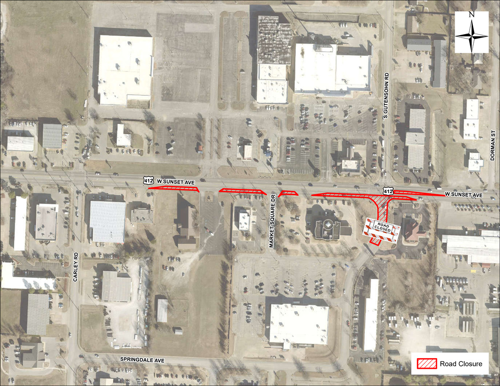 Aerial road closure exhibit depicting the planned construction area at the intersection of West Sunset Avenue (Highway 412) and South Gutensohn Road in Springdale, Arkansas. Highway 412 extends horizontally across the image, while South Gutensohn Road runs north and south near the center-right side of the map. Red shaded and hatched overlays highlight roadway sections affected by the sewer improvement project.  The most significant closure occurs on South Gutensohn Road immediately south of Highway 412, where a large “Road Closed” barricade symbol is placed across the roadway. Additional red-highlighted sections appear along portions of Highway 412 near Market Square Drive and the intersection with South Gutensohn Road, indicating areas where lane restrictions or temporary traffic control measures will be in effect.  Commercial buildings, parking lots, and business entrances surround the intersection. The map includes labels for nearby streets including Carley Road, Market Square Drive, Dorman Street, and Springdale Avenue. A north arrow appears in the upper-right corner to provide orientation, and a legend in the lower-right corner identifies the red hatched markings as roadway closure areas. The exhibit visually communicates the limits of construction activities and the portions of roadway that will be temporarily inaccessible during the utility improvement project.