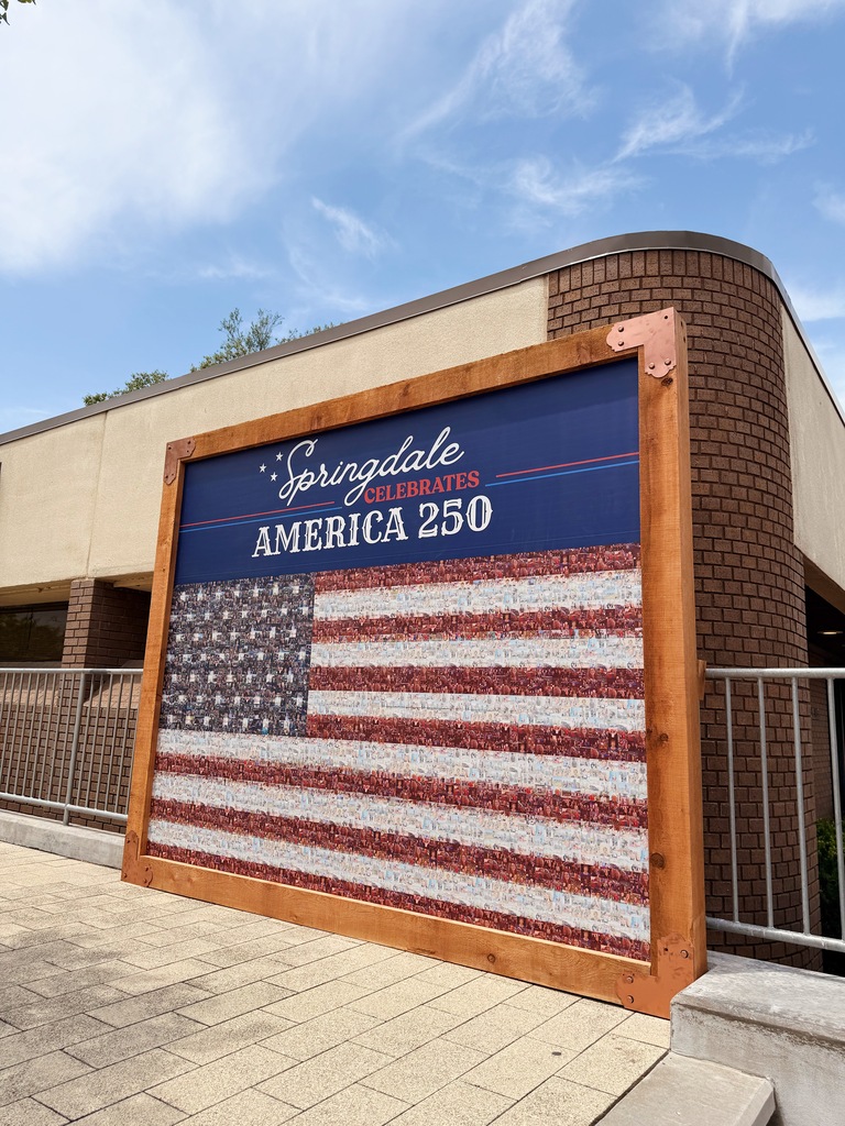 A wide photograph of the "Springdale Celebrates America 250" community flag mural displayed outside along Emma Avenue. The mural consists of an American flag made from hundreds of individual community photographs framed in stained wood with decorative copper corner brackets. Above the flag, a navy-blue panel displays the title "Springdale Celebrates America 250." The mural is positioned beside a tan and brown brick building with metal railings and concrete steps. Overhead, a blue sky filled with soft, wispy clouds provides a bright backdrop, drawing attention to the patriotic display and its prominent downtown location.