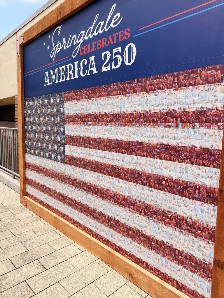 A close-up angled view of the "Springdale Celebrates America 250" community mural highlighting the intricate photo mosaic that forms the American flag. Individual photographs of residents, families, children, veterans, community events, and local scenes are visible throughout the stars and stripes. The navy-blue header at the top features the event title in white and red lettering. The stained wooden frame surrounds the display, while the paved walkway and building exterior are partially visible along the edges of the image. The photograph emphasizes the many individual faces and moments that together create the larger American flag.