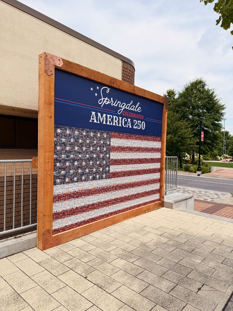 An angled side view of the "Springdale Celebrates America 250" community flag mural installed along the Razorback Greenway on Emma Avenue in downtown Springdale. The large wooden-framed display features an American flag made entirely from hundreds of community-submitted photographs beneath a navy-blue title panel. The mural stands beside a metal safety railing on a light-colored paved plaza. Trees, decorative street banners, and a portion of Emma Avenue are visible in the background, showing the mural's location within the downtown streetscape.