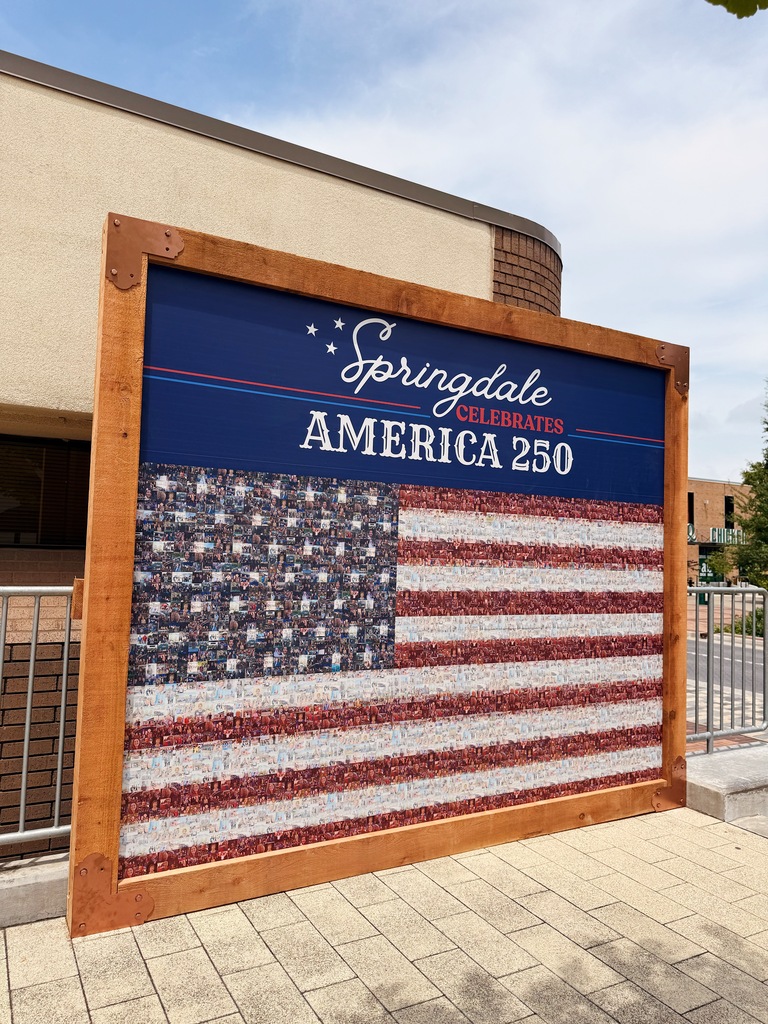 A front three-quarter view of the "Springdale Celebrates America 250" community flag mural. The large American flag mosaic, created from hundreds of photographs submitted by local residents, fills most of the display beneath a navy-blue title panel reading "Springdale Celebrates America 250." The mural is enclosed within a stained wooden frame with decorative copper corner plates. A brick commercial building and part of Emma Avenue can be seen in the background, while the paved plaza in front of the mural provides space for visitors to stop, view the artwork, and search for their own photographs.
