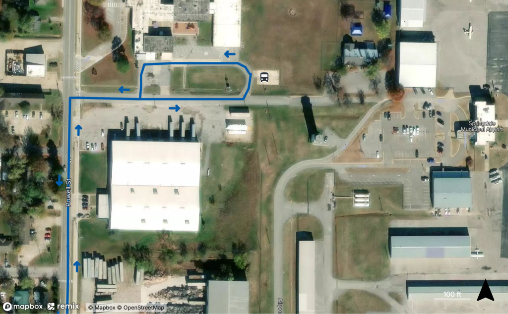 An aerial map showing the second designated shuttle parking location for the Helos, Wings & Wheels event near Jones Elementary. Blue arrows illustrate the recommended traffic pattern entering from Powell Street, continuing east along the access road into the parking lot, and exiting back toward Powell Street. A shuttle pickup location is identified with a bus icon near the parking area. The map provides visitors with directions for using overflow parking and accessing the free shuttle service to the Springdale Municipal Airport.