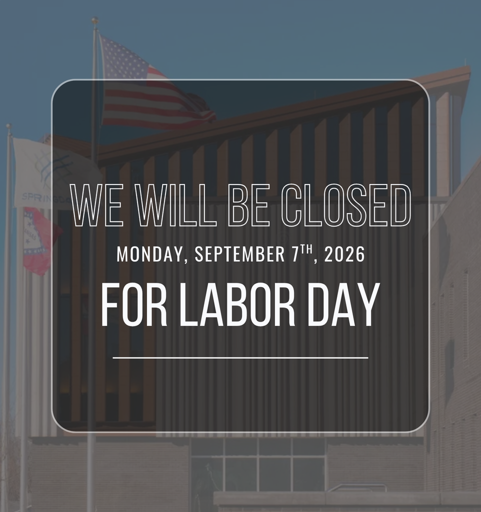 A Labor Day closure announcement graphic featuring a full-background photograph of Springdale City Hall. The image shows the front exterior of the multi-story brick municipal building, with tall, narrow vertical windows and architectural columns extending across the center. A large American flag is prominently displayed on a flagpole in the upper left portion of the image, with additional flagpoles and partially visible flags along the left side of the building. The photograph has a muted blue-gray overlay that darkens the building and flags so the announcement text is easy to read. Centered over the photograph is a large, dark, semi-transparent rectangular text box with rounded corners and a thin light-gray border. Inside the box, large outlined white uppercase letters read, “WE WILL BE CLOSED.” Directly underneath, smaller white uppercase text reads, “MONDAY, SEPTEMBER 7TH, 2026.” Below that, very large bold white uppercase letters read, “FOR LABOR DAY.” A thin horizontal white line appears beneath the text near the bottom of the box. The overall design uses a subdued, professional color palette of dark gray, blue-gray, white, and the muted red, white, and blue of the American flag.