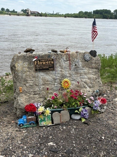 Large stone with memorial plaque and flowers on and around it with the Mississippi River in the background