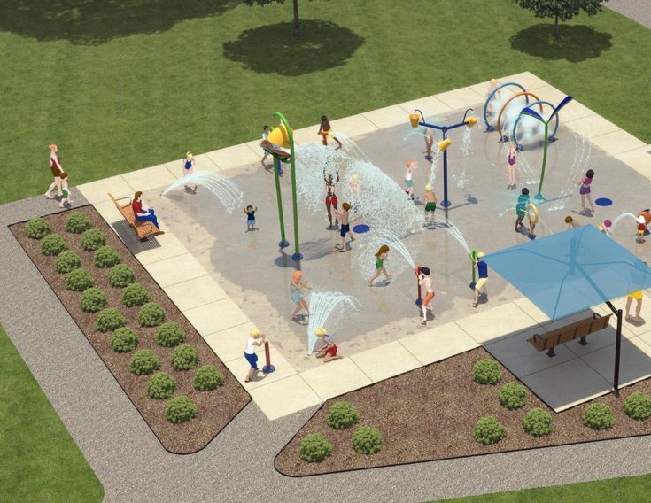 Splash pad with bucket and spray features with fencing surrounding the splash pad and trees and green space in the background