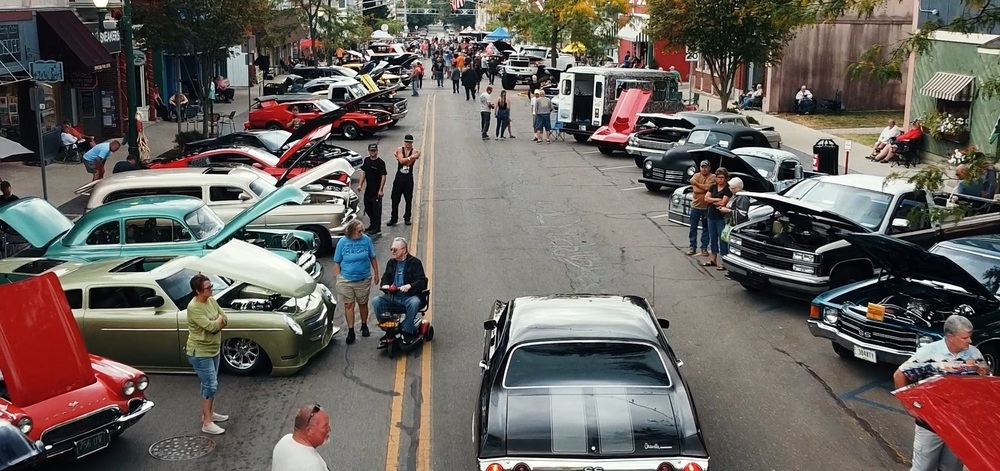 Downtown street filled with classic cars parked along both sides, many with hoods open for display, as community members walk, talk, and admire vehicles during a lively cruise-in event.