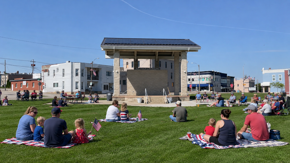 Families gather on picnic blankets with small American flags on a sunny lawn in front of the Artisan Crossing pavilion during a small-town community event.