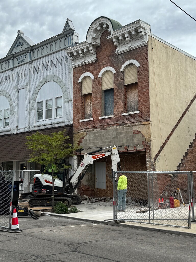 Historic brick building under construction with excavator, fenced work area, and a worker in safety gear on a downtown street.