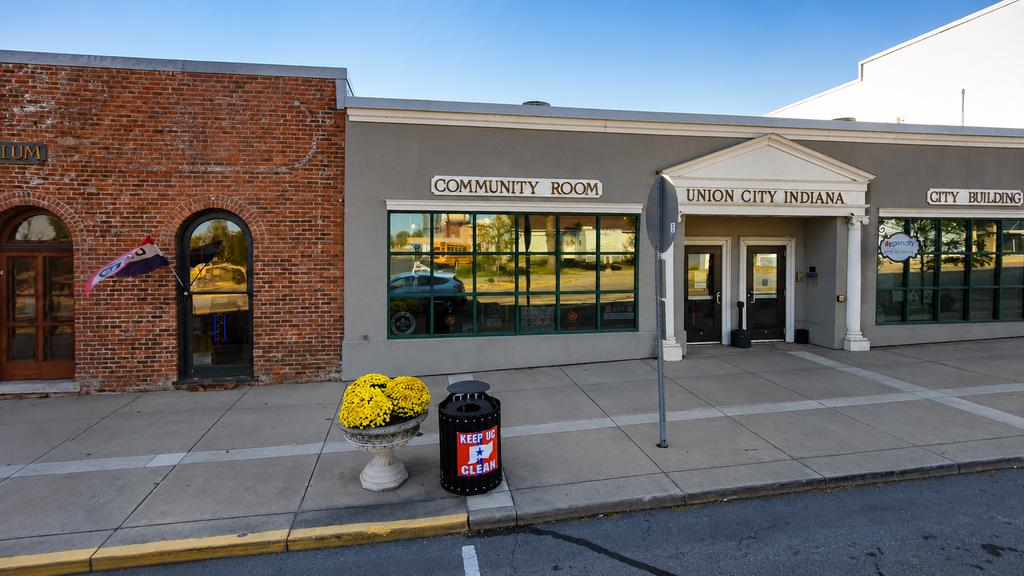 City building exterior with “Community Room” and “Union City Indiana” signage, large front windows, and a sidewalk with a planter and trash can in front.
