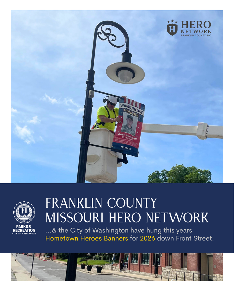 a man in a cherry picker putting a Hometown Heroes Banner on the light post down Front Street. Reads: Franklin County Missouri Hero Network & the City of Washington have hung this years Homwtown Heros Banners for 2026 down Front Street.