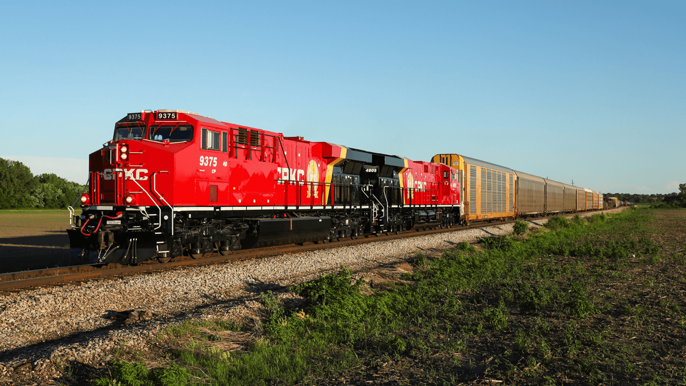 Red train engine with the letters "CPKC" on the side in white text. The engine is hauling cars through the countryside