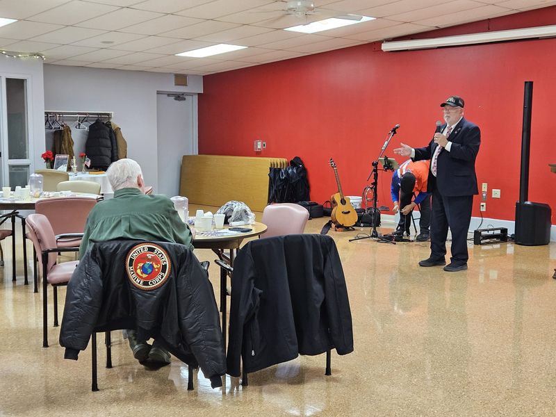 Paul Hieser speaking into a microphone in front of a group of Veterans