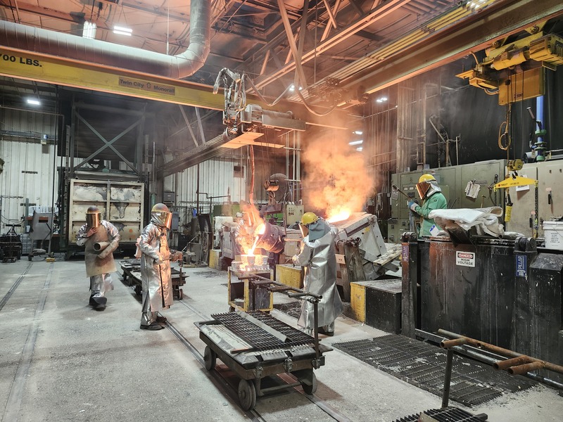 People in protective clothing make machine pieces from moltem metal in factory setting.