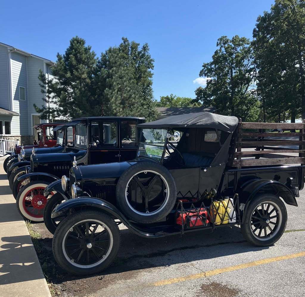 Three Ford Model T's lined up in front of a sidewalk, with blue skies and tall trees behind them.