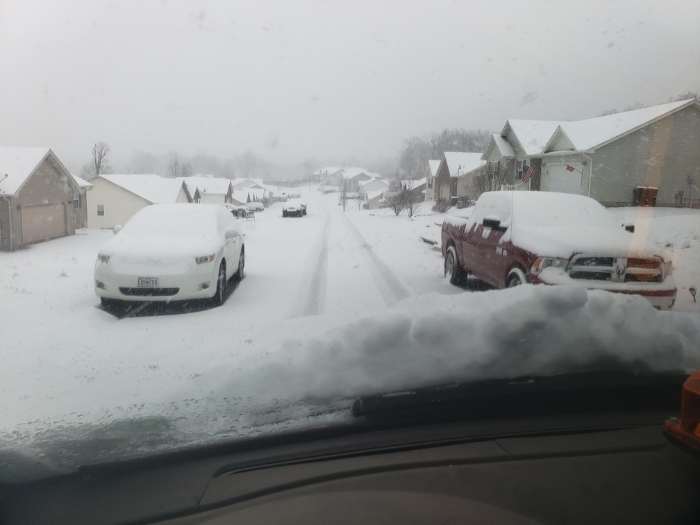 Roadway with cars double parked, covered in snow; snowplow cannot clear roadway due to vehicles. 