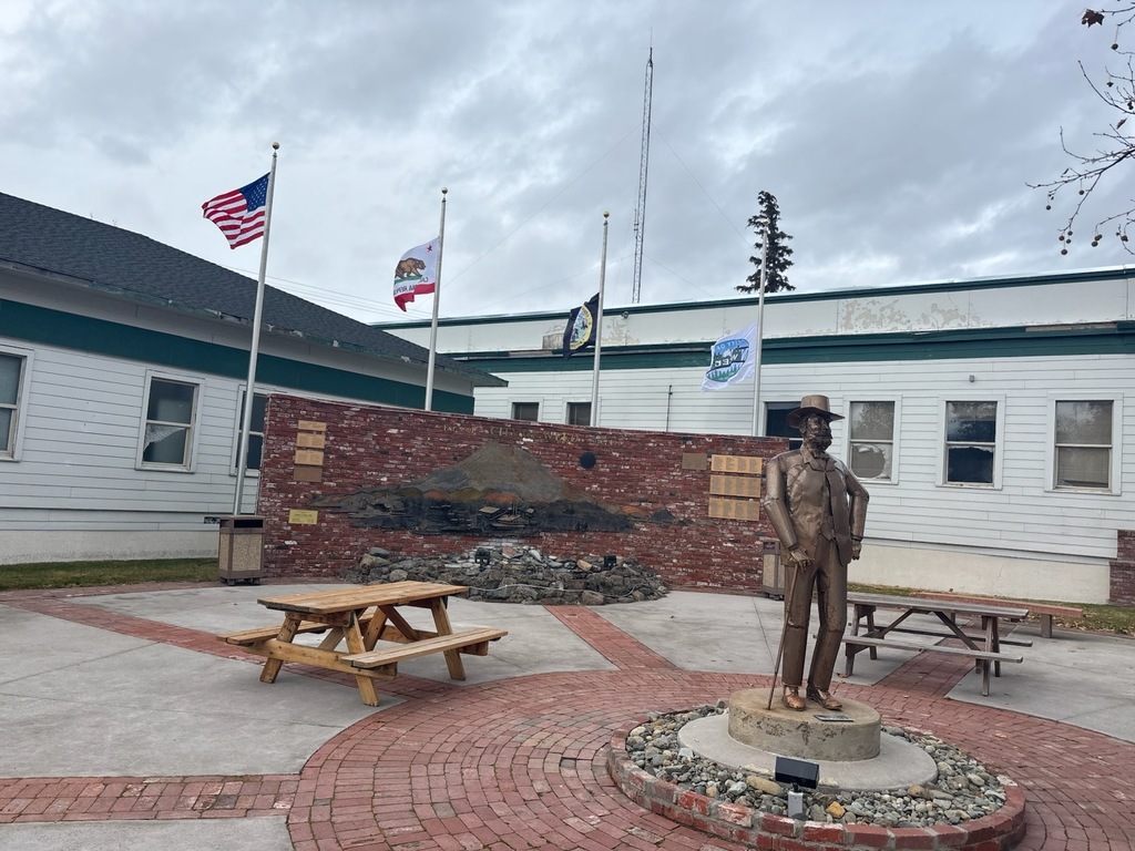 City park courtyard featuring four flagpoles with the U.S. flag, California state flag, city flag, and an additional commemorative flag flying behind a statue of Abner Weed, the city’s founder. Picnic tables and brick walkways surround the monument, with City Hall buildings in the background under an overcast sky.