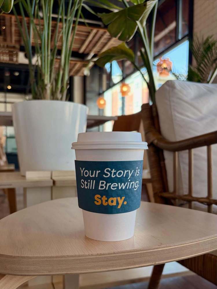 A take-away coffee cup with a sleeve that says "Your Story is Still Brewing. Stay."