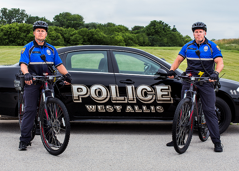A photo of a West Allis Police Department car with two officers on bicycles posing in front of the car.