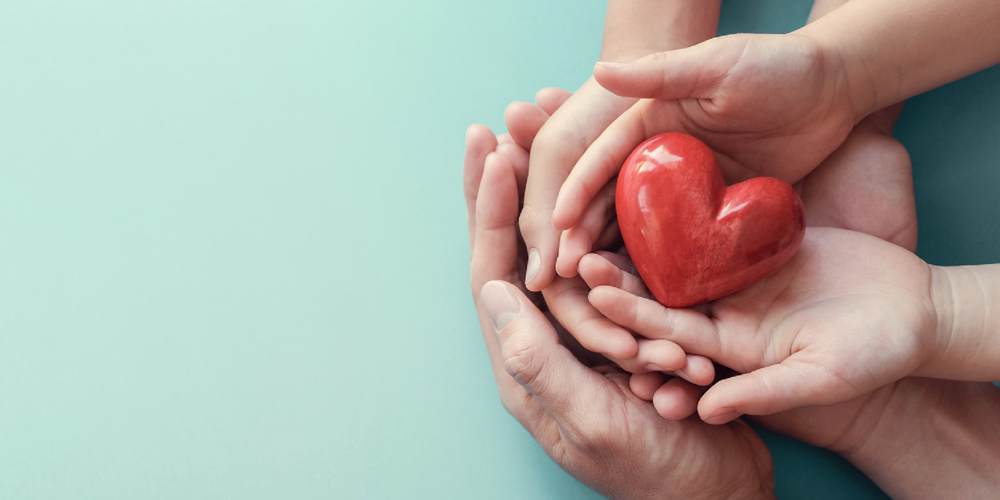 Three sets of hands on top of each other facing palms up holding a heart figurine