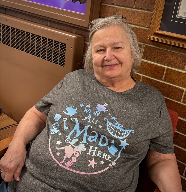 Woman with gray hair sitting on a chair smiling at the cameras, wearing a "We're all mad here" t-shirt. 
